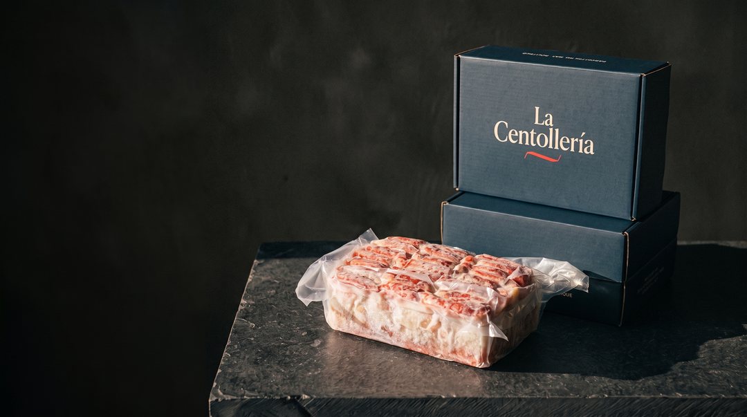 Bloque congelado de carne de centolla en su bolsa al vacío sobre una mesa de piedra oscura; atrás, dos cajas navy de La Centollería apiladas