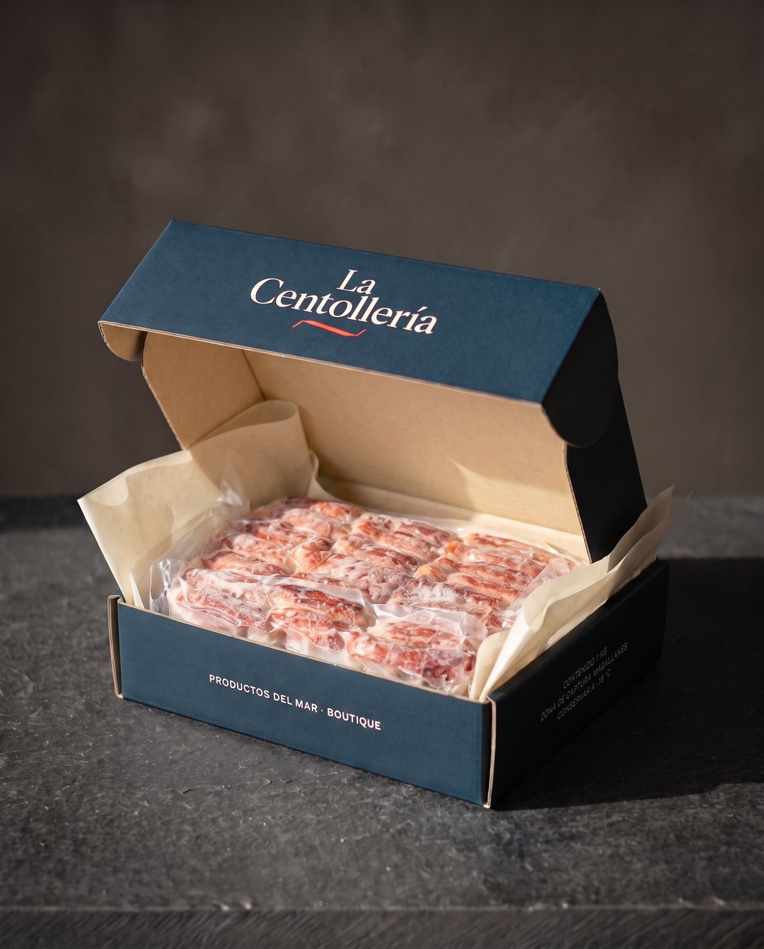 Caja navy de La Centollería abierta, con interior kraft y papel de seda, y adentro el bloque congelado de carne de centolla en su bolsa al vacío, sobre una mesa de pizarra