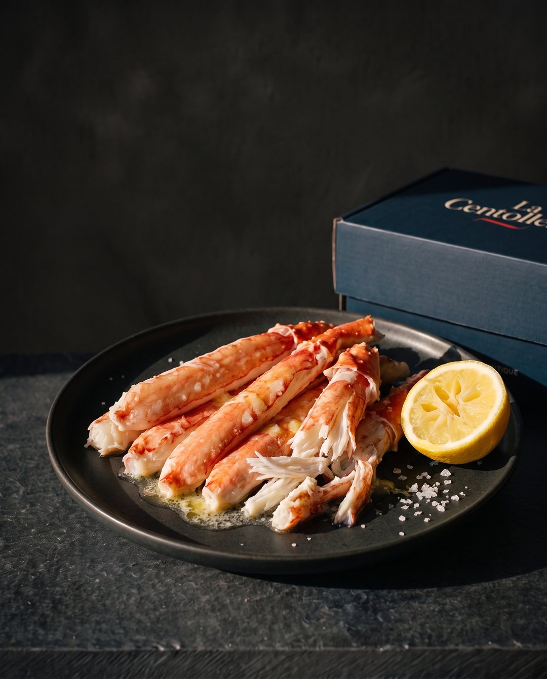 Plato de carne de centolla tibia, sin caparazón, con mantequilla derretida, sal gruesa y medio limón sobre una mesa de pizarra; al lado, la caja navy de La Centollería, con sol bajo de tarde